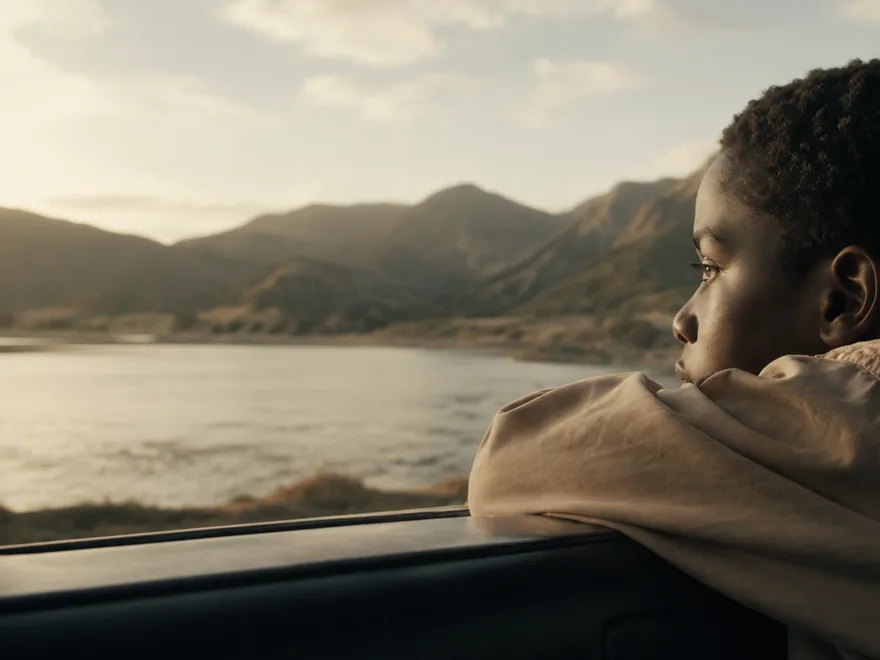 A child resting on a car window, watching a mountain lake at sunset.